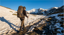 Escursionista con zaino da trekking su sentiero di montagna tra neve residua e terreno roccioso in primavera